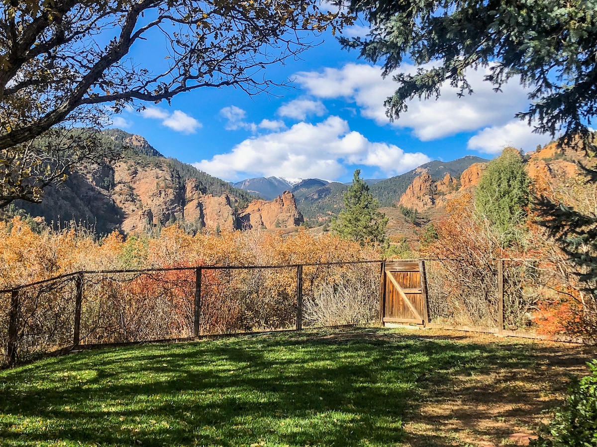 A fenced backyard offers a view of the surrounding mountains and autumn foliage. The grassy area is framed by trees, with a wooden gate visible, leading out to nature trails. The sky is partly cloudy, adding depth to the scenic landscape.