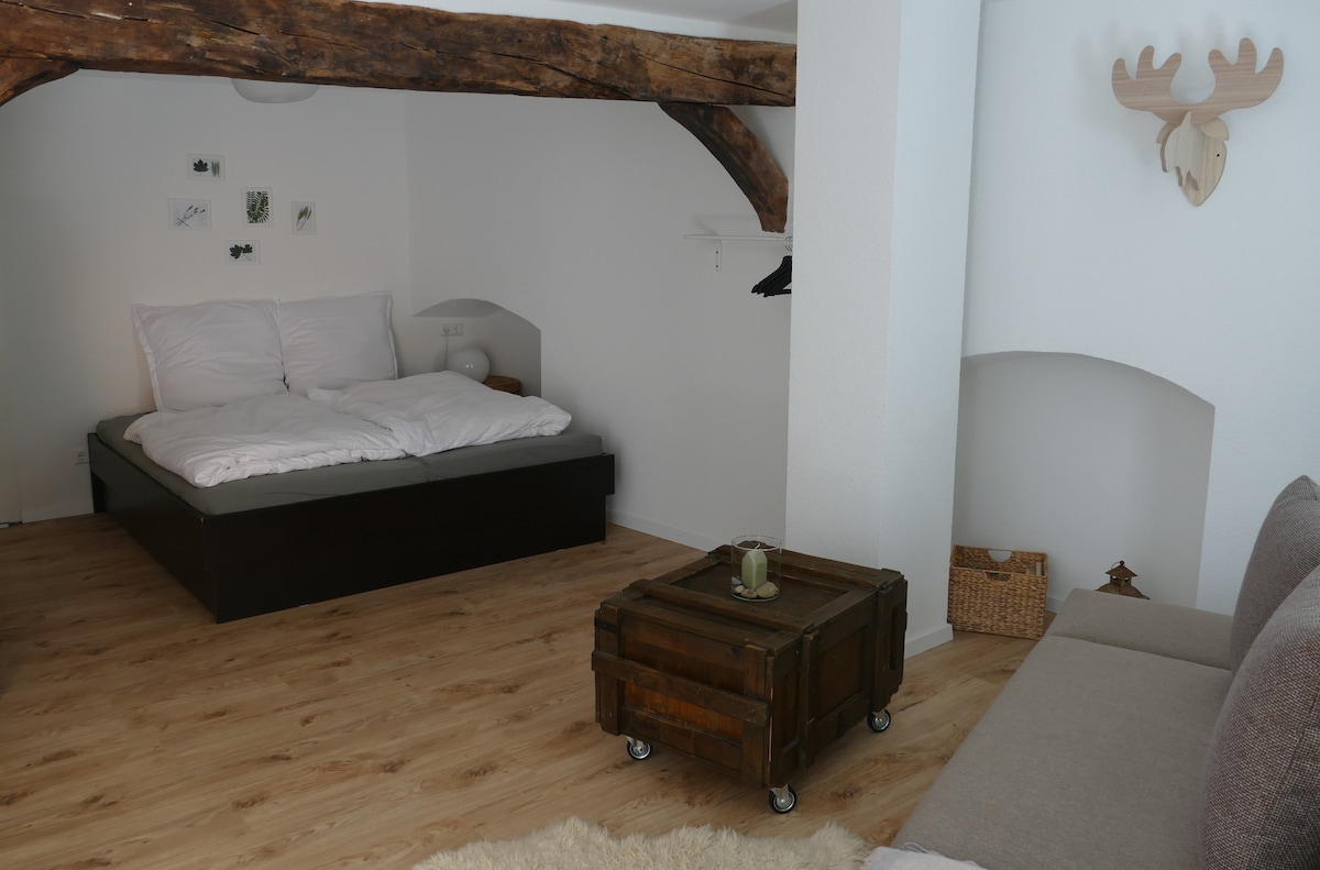 The open sleeping area features a spacious bed with crisp white linens, positioned under a rustic wooden beam. A cozy seating area is provided with a light sofa, and a wooden trunk serves as a coffee table. Minimalist decor enhances the simplicity of the space.