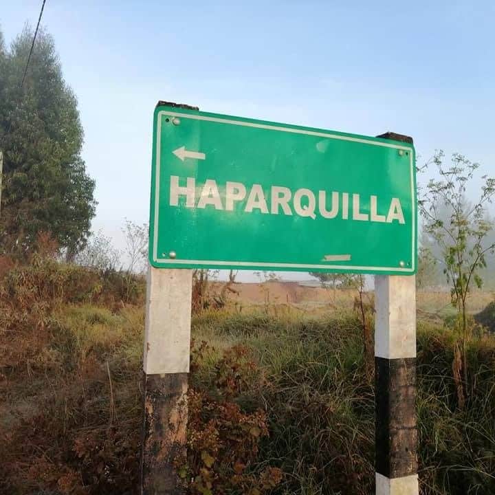 A green roadside sign displays the name 'Haparquilla' in large white letters. The sign is supported by a black and white striped pole and is surrounded by lush greenery. A soft mist is present in the background, enhancing the tranquil atmosphere.