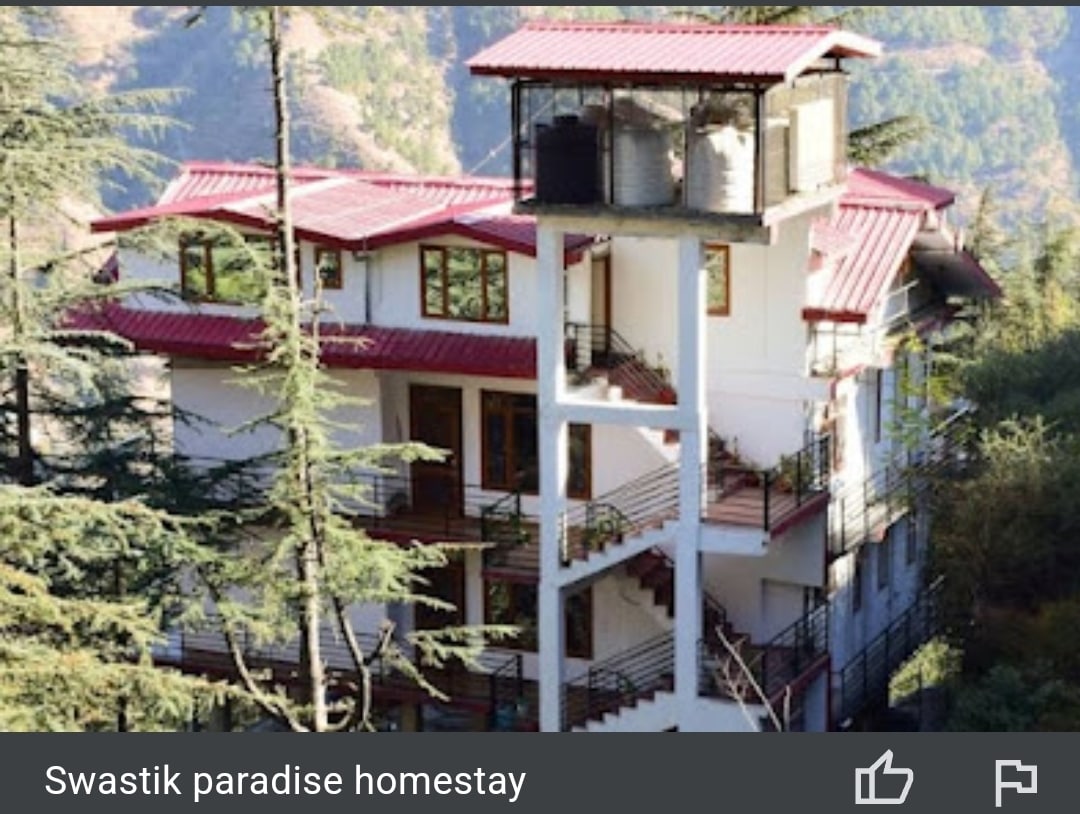 The multilevel homestay features a modern exterior with a red metal roof and white walls. Large windows provide ample natural light, while a balcony extends from the second floor, framed by surrounding trees and natural elements. A staircase leads to the entrance.