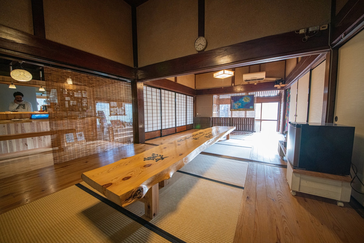 A spacious common area features a 4-meter long wooden table at its center, set on tatami flooring. Natural light filters through shoji screens, illuminating the room's warm wooden accents. A counter bar is visible in the background, complementing the inviting layout.