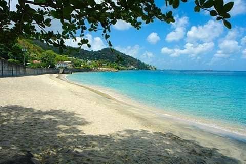 A sandy beach stretches along calm turquoise waters, bordered by lush greenery and gentle hills in the background. A clear blue sky is filled with soft, white clouds, providing a serene coastal atmosphere.