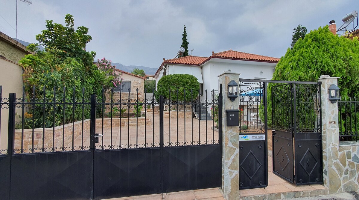 An inviting exterior of Tyros House is displayed, featuring a secure black gate with decorative elements. Lush greenery and well-maintained gardens are visible alongside paved pathways. The structure's white walls and terracotta roof add charm to the inviting entrance, surrounded by a peaceful atmosphere.