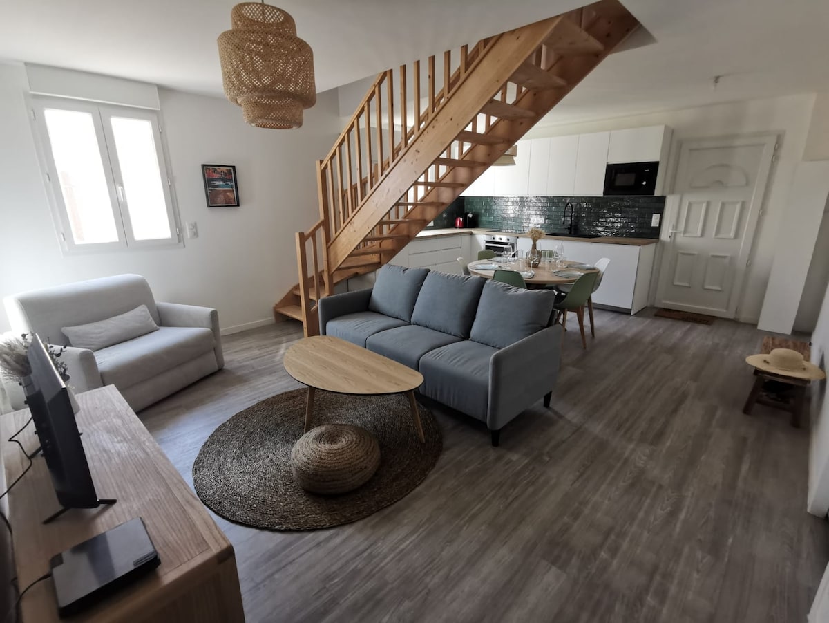 A modern living space features a light-colored sofa and a round coffee table on a textured rug. The open kitchen is visible, highlighted by white cabinetry and a dark backsplash. Natural light enters through two windows, while a wooden staircase leads to the upper level.