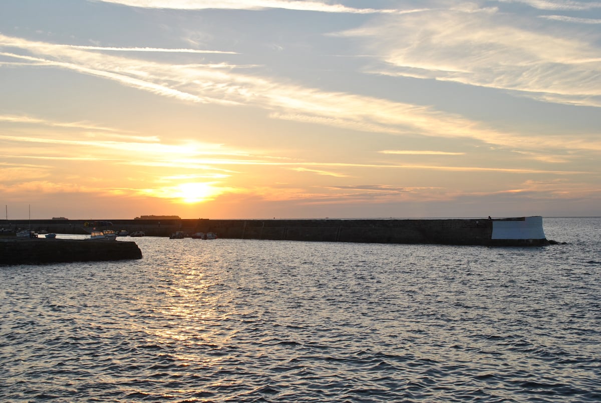 A view of the harbor at sunset, where gentle waves reflect golden sunlight. A stone pier extends into the water, while silhouettes of distant structures can be seen against the colorful sky, which displays soft shades of orange and blue.