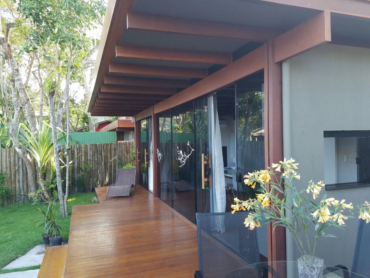 A spacious outdoor deck is framed by large glass doors, providing a seamless connection to the interior. Lush greenery surrounds the area, enhancing the natural setting. A table with chairs and a decorative vase of flowers adds a touch of detail to the space.