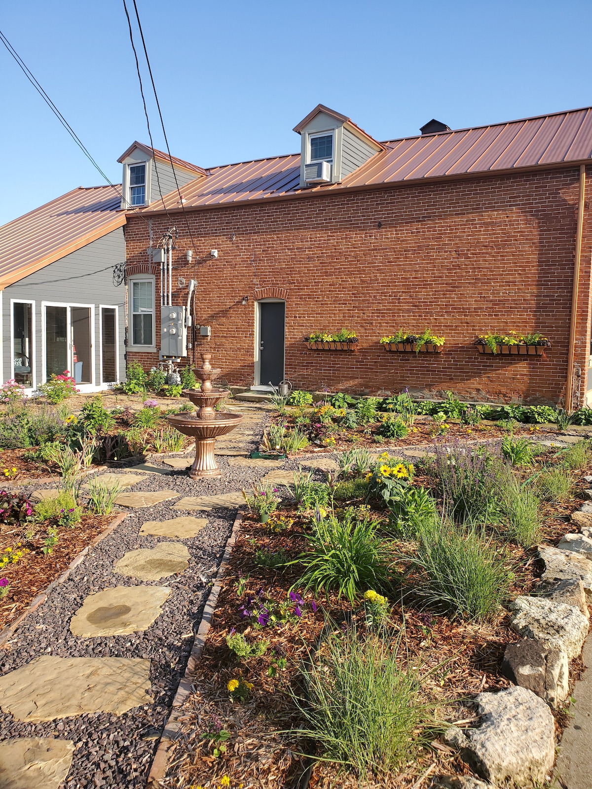 A charming outdoor pathway is lined with lush garden beds and colorful flowers, leading to a brick building with a modern metal roof. A decorative fountain sits centrally, enhancing the tranquil atmosphere. Wall-mounted planters add greenery to the structure, contributing to the inviting landscape.