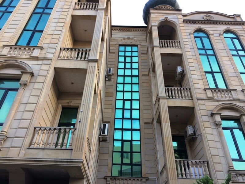 The exterior of a villa is shown, featuring a harmonious blend of classic architecture and modern glass elements. Tall windows reflect the surrounding environment, while ornate balconies add character to the façade. The stone detailing enhances the building's elegant appearance.