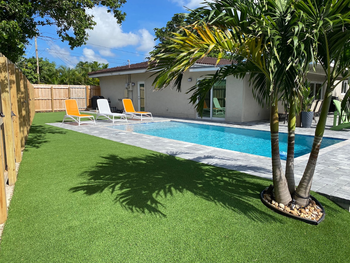 A heated pool is surrounded by meticulously maintained landscaping, featuring palm trees and artificial grass. Sun loungers are positioned near the pool area, providing a relaxed space for sunbathing. The exterior of the house offers large windows that allow natural light to enter.