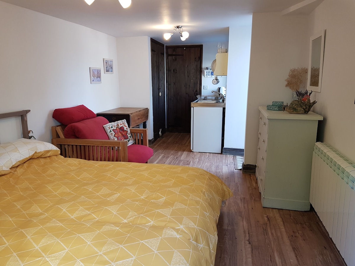 A spacious room features a comfortable king-size bed covered with a textured yellow quilt. A kitchenette is visible in the background, alongside a wooden table. Natural light enters from a nearby window, illuminating the simple decor, which includes a dresser and casual seating.