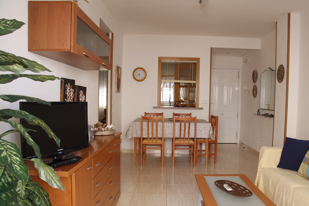 A light-filled living area features a wooden dining table set for four and a wooden TV stand with a television. A large plant adds greenery to the space, while soft furnishings in neutral tones create a comfortable atmosphere. A wall clock and decorative items enhance the decor.