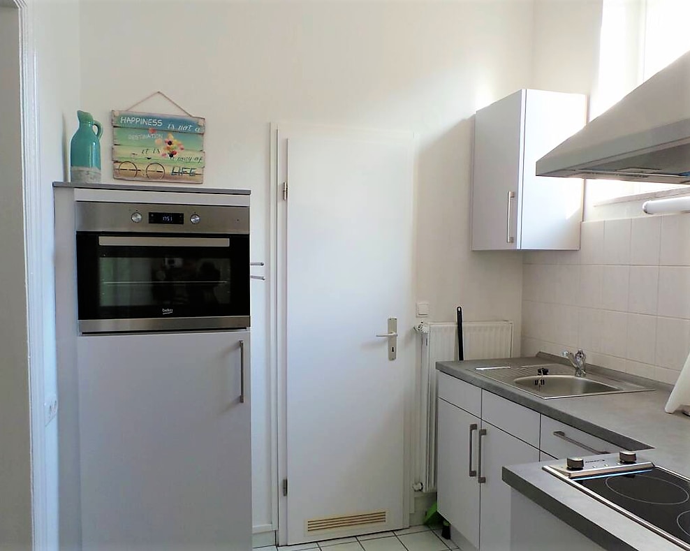 A compact kitchen area is depicted, featuring modern appliances including an oven, refrigerator, and stovetop. Light-colored cabinets and a clean countertop contribute to a fresh ambiance. Natural light enters through a nearby window, enhancing the functional space.