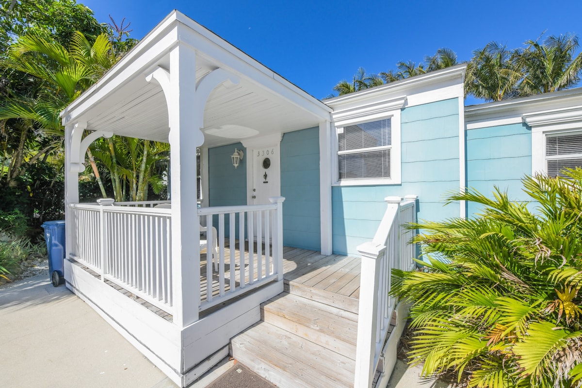 A charming beach cottage entrance showcases a covered porch with white railings and steps leading up to the door. Lush tropical plants frame the path, while blue siding reflects a coastal vibe under a clear blue sky.