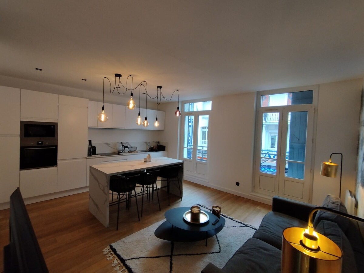 A modern kitchen and living area feature sleek cabinetry and counter space. The room is illuminated by elegant pendant lights. A comfortable sofa and round coffee table are present, with large windows allowing natural light to enter and a view of the outdoors.