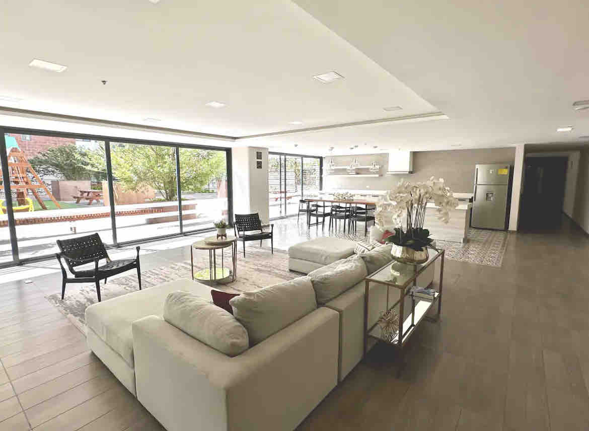 A spacious living area is presented with a cozy sectional sofa and modern coffee table. Large glass doors allow natural light to fill the space and provide a view of the outdoor play area. A dining table is visible in the background, suitable for group gatherings.