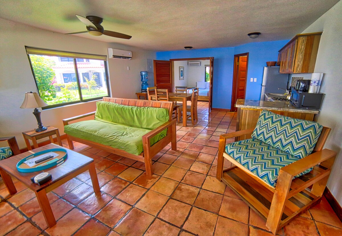 An open-plan living area is furnished with a green futon sofa and a patterned armchair. A dining table is visible alongside a fully equipped kitchen. Large windows allow natural light to fill the space, showcasing terracotta tile flooring and vibrant wall colors.