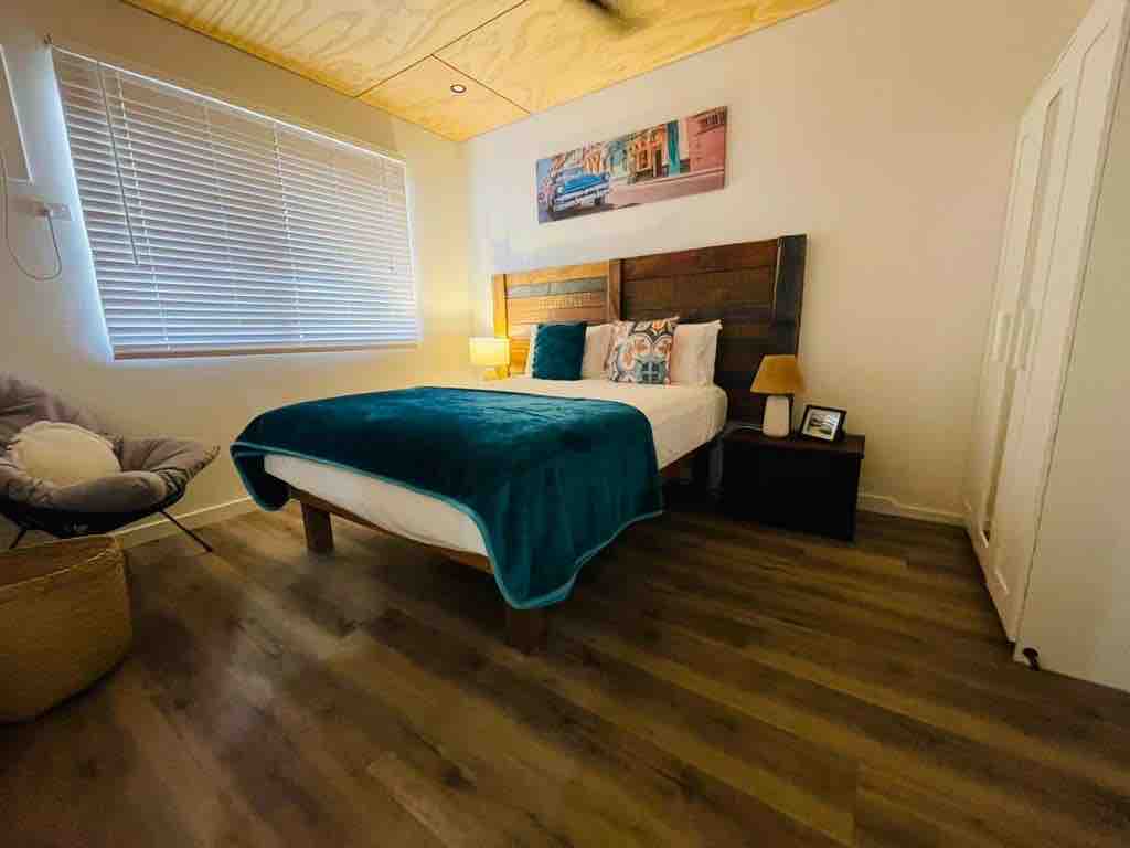 A well-appointed bedroom features a plush queen bed with a turquoise throw blanket. A wooden headboard is complemented by decorative pillows. Soft lighting is provided by two bedside lamps. A cozy chair sits in the corner, while a wardrobe is positioned nearby beneath a window with blinds.