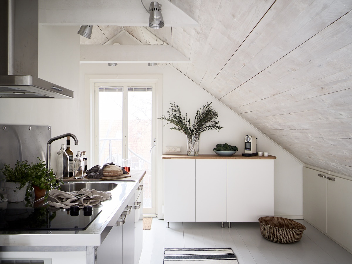 A modern kitchen features vaulted wooden ceilings and sleek cabinetry. A stove and sink are positioned alongside a countertop, with decorative plants placed nearby. Natural light floods the space through double doors, leading to a balcony. A woven basket sits on the floor.