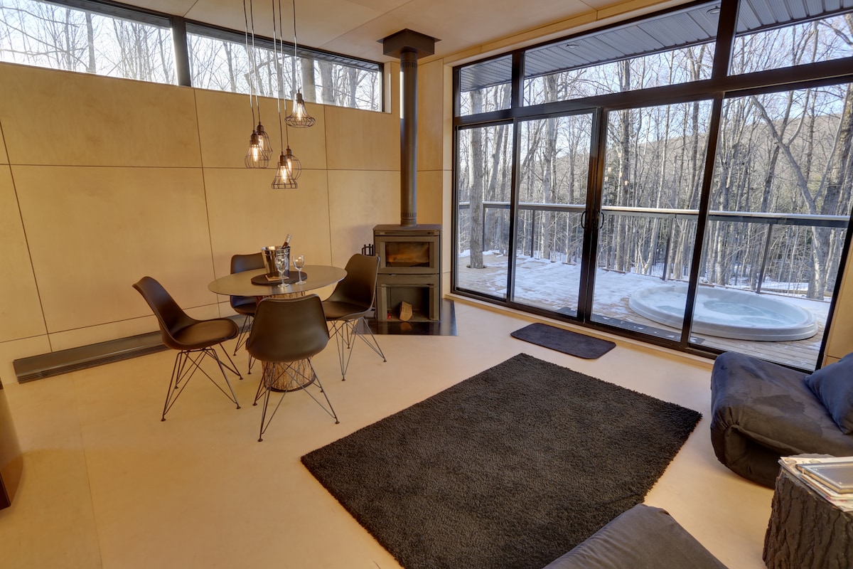 A modern dining area features a round table surrounded by four sleek chairs, with a large window offering views of the snowy landscape outside. A cozy seating area is placed nearby, while a glass door leads to a private deck with a hot tub.