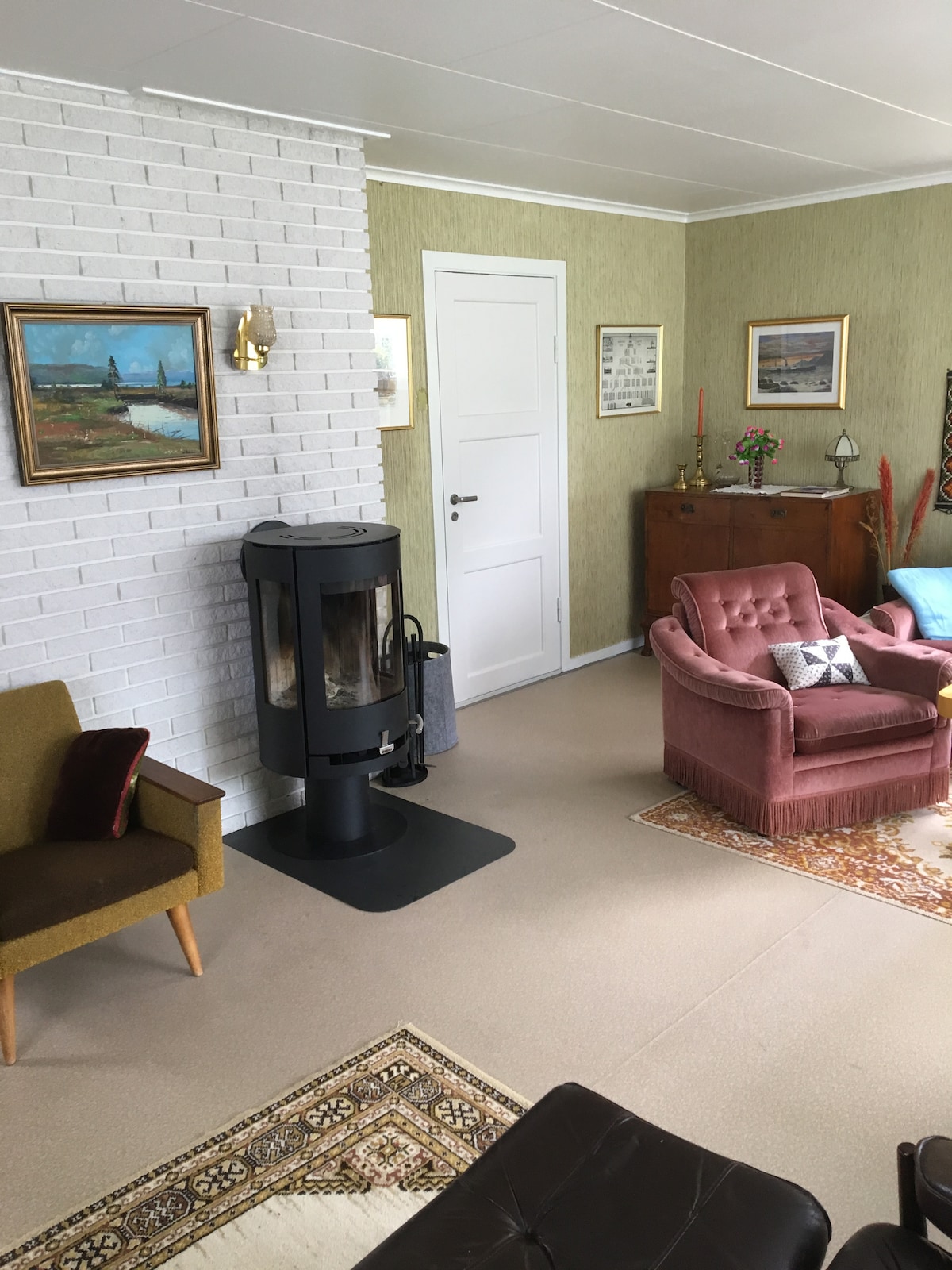 A comfortable living area is highlighted by a decorative fireplace, with various artworks adorning the walls. Furniture includes a pink armchair, a green chair, and a vintage wooden cabinet, complemented by a patterned rug on the floor.