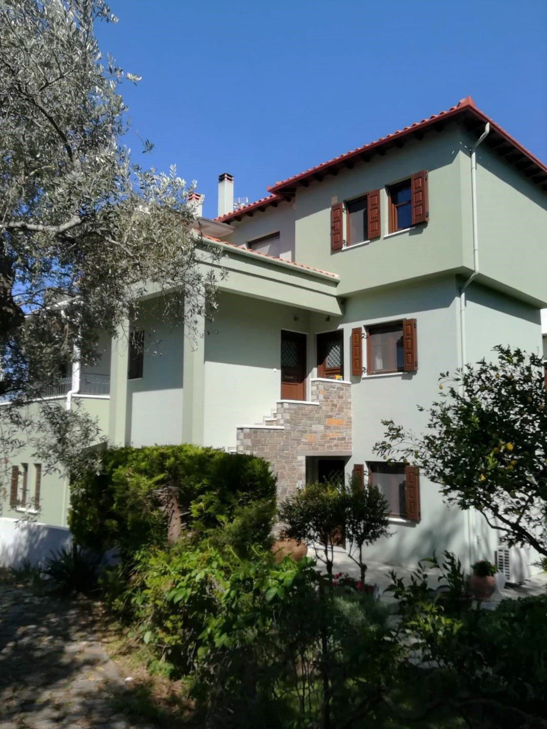 The exterior of the modern home is framed by lush greenery and decorative plants. Large windows with wooden shutters enhance the façade, while a stone pathway leads to the entrance. The property features multiple levels, with a charming red-tiled roof contributing to its inviting appearance.