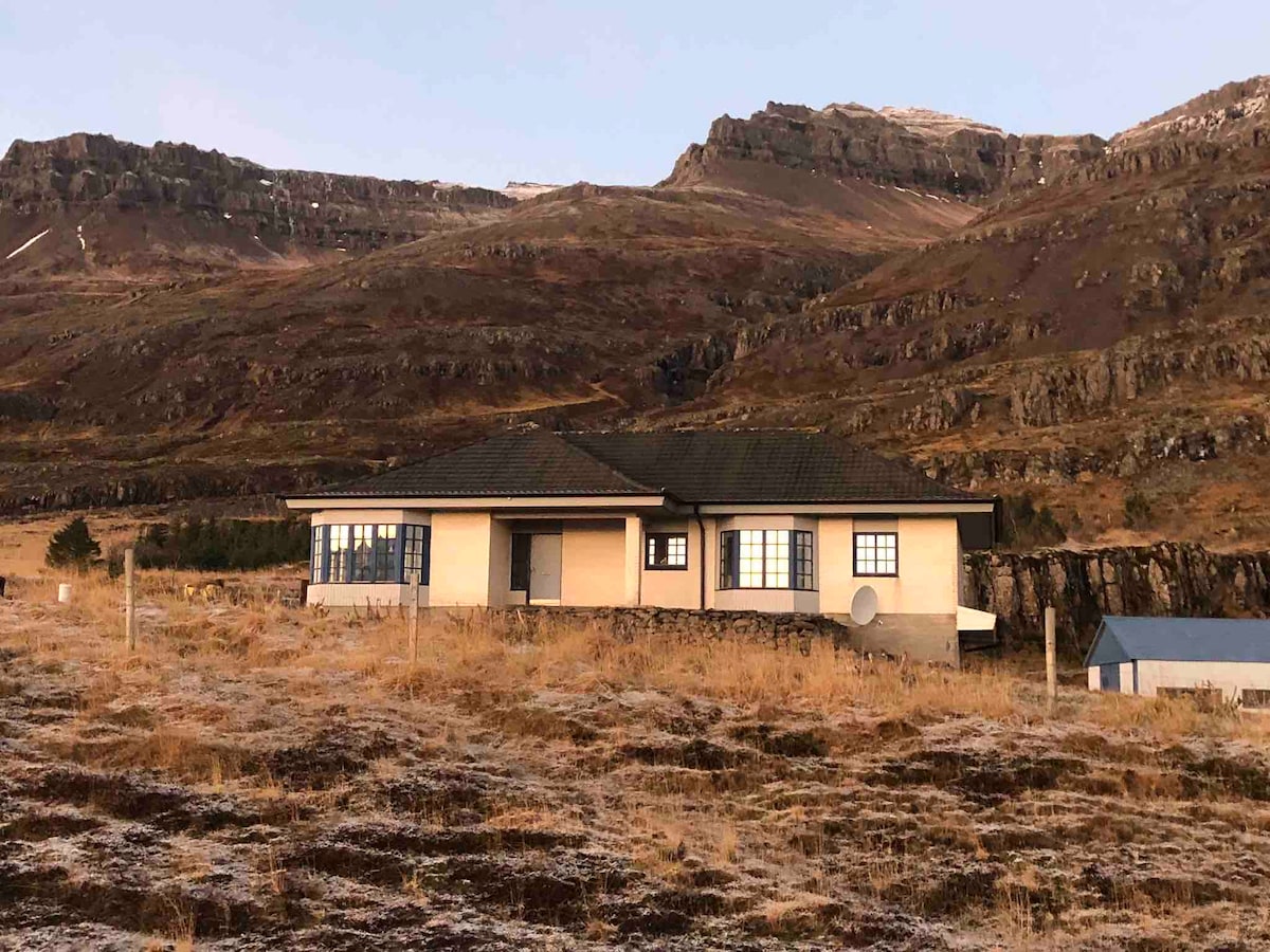 The house is located in a natural setting, surrounded by gently rolling hills. The two-story structure features large windows that allow plenty of natural light. An expansive view of the landscape is complemented by the rugged mountains in the background, creating a serene environment.