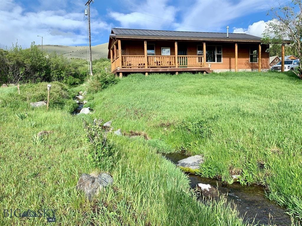 The cozy cabin, built from warm wood, is surrounded by lush green grass and a gentle creek flowing nearby. A covered deck extends from the front, offering a space to enjoy the outdoors. Scenic mountains can be seen in the background under a partly cloudy sky.