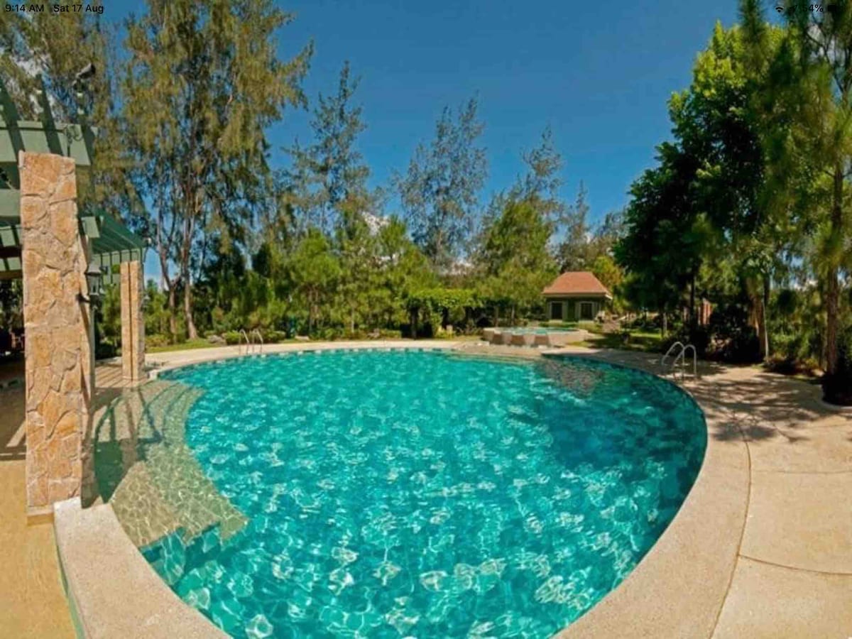A large swimming pool is surrounded by lush greenery and trees, with clear blue water reflecting the sky. A shaded area with a stone structure is visible nearby, providing a relaxing space. The setting offers a serene and inviting atmosphere for guests.
