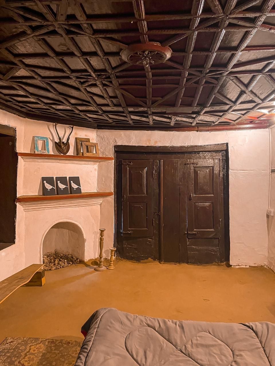 A corner of the room showcases a simple, traditional fireplace beneath a decorative wooden shelf. Two framed artworks are displayed on the shelf, while double wooden doors provide access to another area. The ceiling features distinctive, intricate wooden detailing.