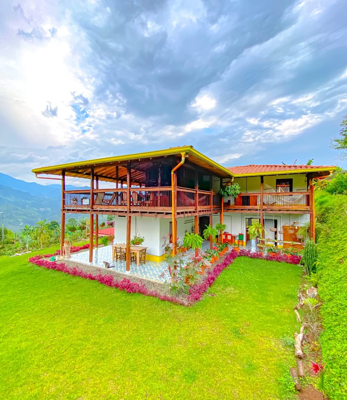 A two-story house is set against a backdrop of rolling hills and cloudy skies. The exterior is complemented by vibrant greenery and a well-maintained garden. Outdoor seating areas with tables and chairs are visible on the terrace, inviting guests to enjoy the scenic surroundings.