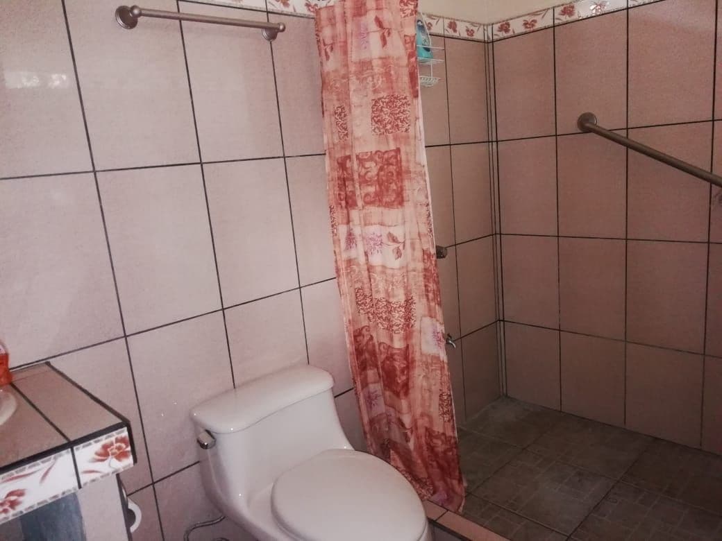 A bathroom featuring tiled walls and floor is shown. A toilet is positioned beside a washbasin with a counter. A shower area with a textured curtain and safety grab bar is visible, providing accessibility. Neutral tones and simple decor enhance the practicality of the space.