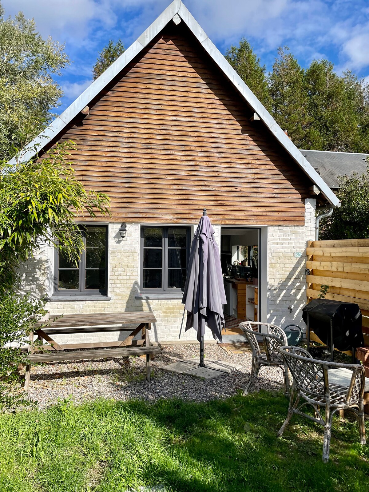 The exterior of a renovated barn features a wooden facade and a triangular roof. A patio area includes a wooden bench and a table with four chairs, complemented by a large umbrella. Lush greenery surrounds the space, creating a serene outdoor environment.