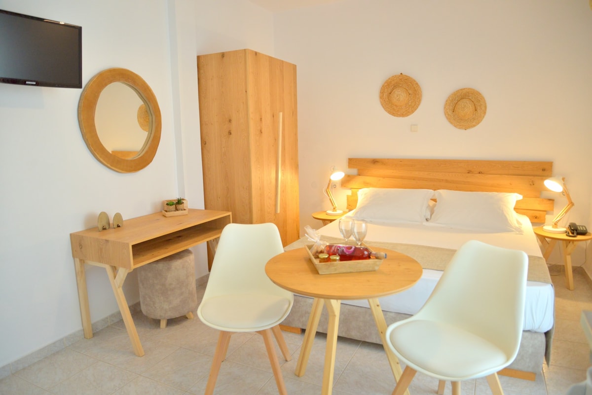 A bright studio is presented with a spacious double bed, complemented by wooden accents. A round table with two chairs is set next to the bed, while a wooden wardrobe and a wall mirror enhance the functional layout. A television is mounted on the wall.