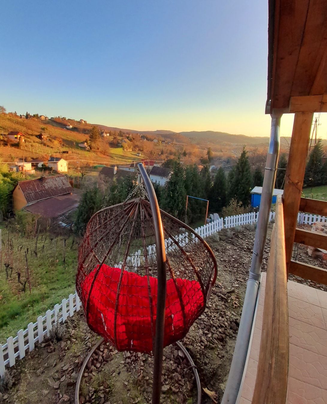 A red hanging chair is positioned on a wooden terrace, offering panoramic views of the rolling hills. The gently sloping landscape is dotted with trees and houses, bathed in warm light from the setting sun. A white picket fence outlines the yard.