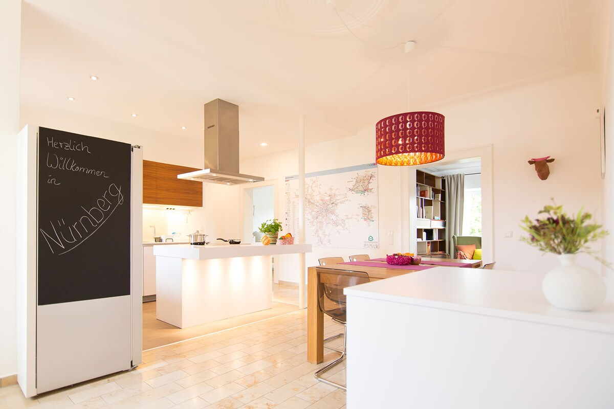 A modern kitchen and dining area is presented, featuring a central island with a sleek countertop. Stylish wooden cabinetry complements the space. A chalkboard stand welcomes guests, while a vibrant light fixture adds a pop of color. Natural light illuminates the room through large windows.