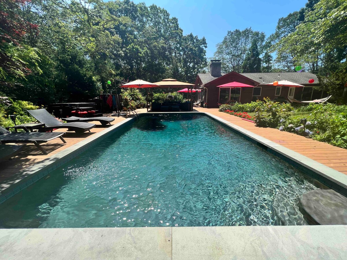 An in-ground saltwater pool is surrounded by wooden decking and lounge chairs, offering a serene outdoor space. Sun umbrellas provide shade, and lush greenery is visible along the perimeter, enhancing the natural ambiance of the property.