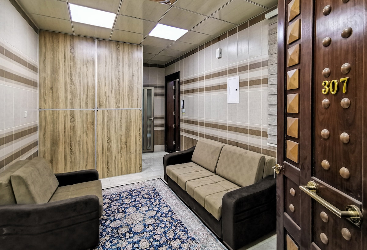 A lobby area features two dark sofas facing each other, complemented by a decorative area rug. A divider with wood paneling adds privacy, and light fixtures illuminate the space. A wooden door with a decorative handle and the room number is visible in the foreground.