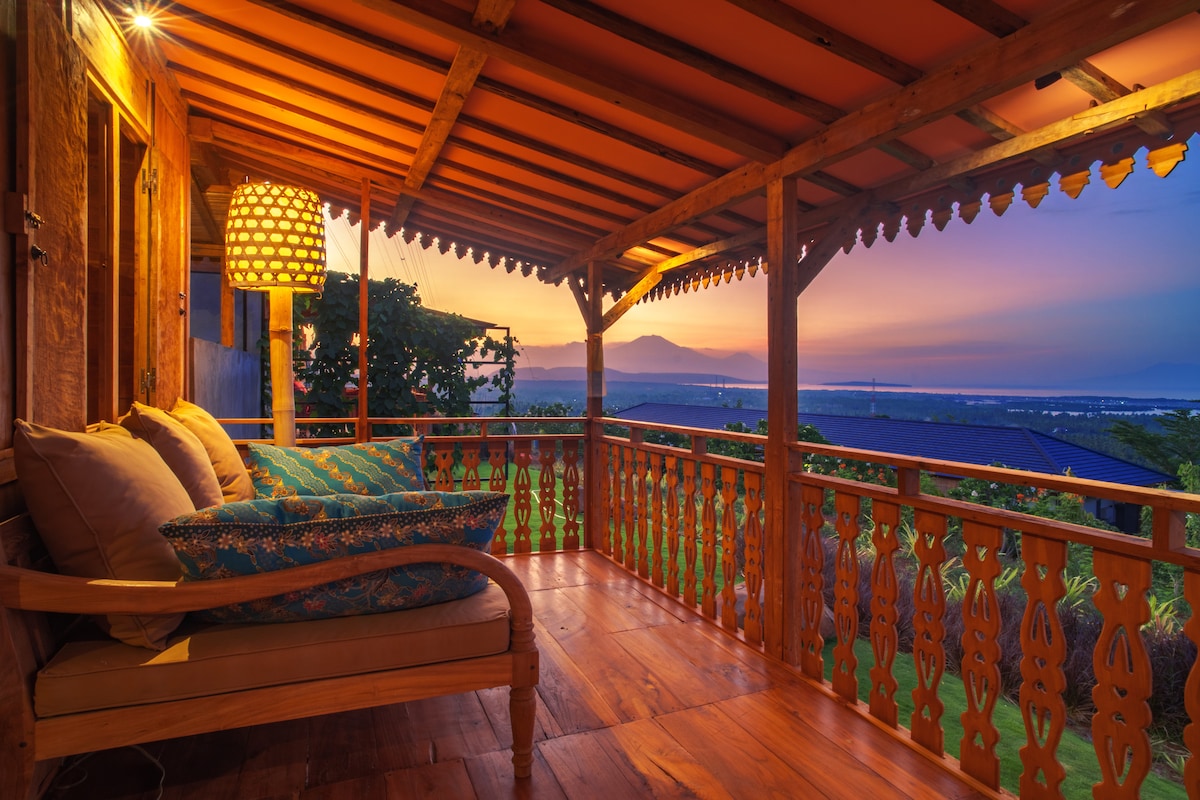 An inviting wooden terrace features a comfortable seating area adorned with plush cushions. The warm glow of a woven lamp illuminates the space, while a breathtaking sunset casts vibrant hues across the sky, enhancing views of the surrounding landscape and distant volcanoes.