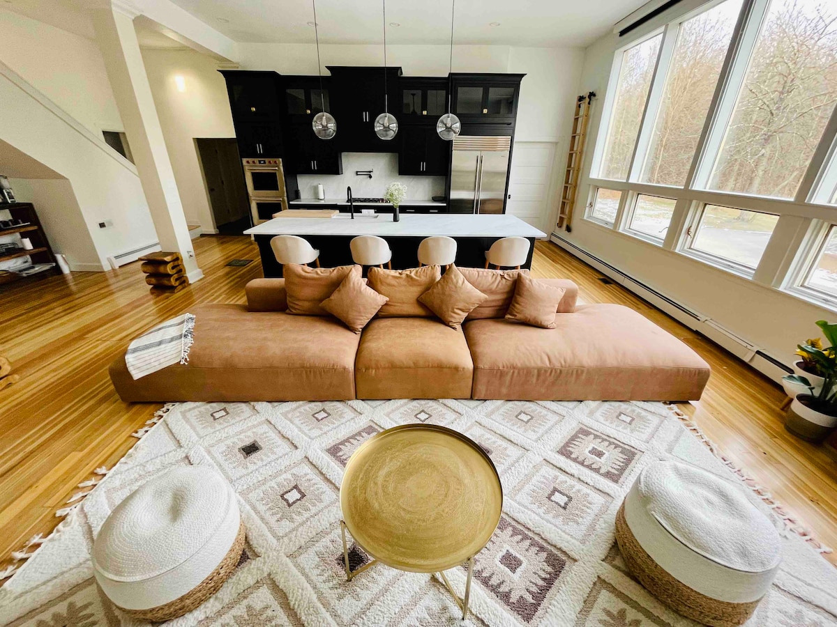 A spacious living area features a wide, low-profile sofa adorned with decorative pillows, facing a sleek kitchen with dark cabinetry. A round coffee table rests on a textured area rug, accompanied by two poufs. Large windows let in natural light, highlighting the warm wood floors.