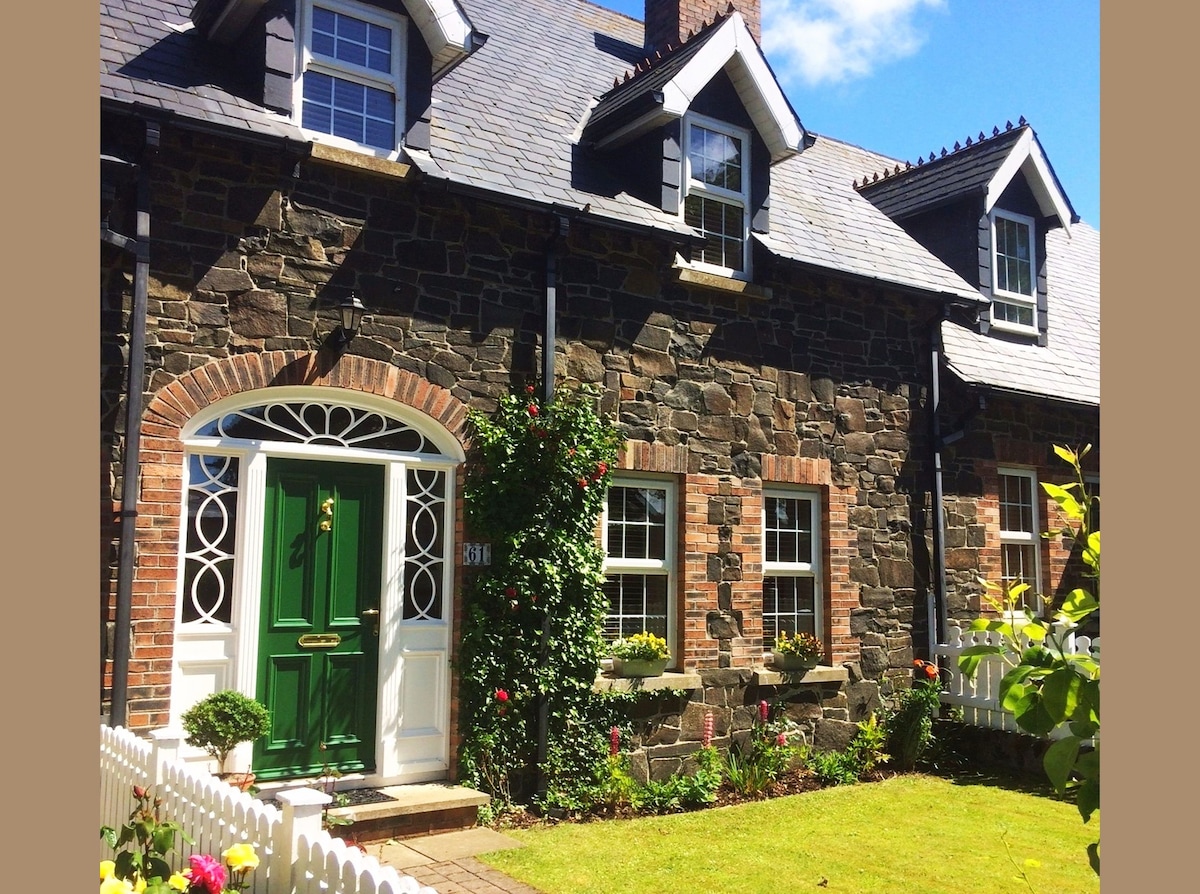 A traditional stone cottage exterior features a vibrant green front door and white trim. Flowerbeds with colorful blooms are positioned alongside the neatly trimmed lawn. Windows with decorative shutters are present, highlighting the charming architectural details of the cottage.