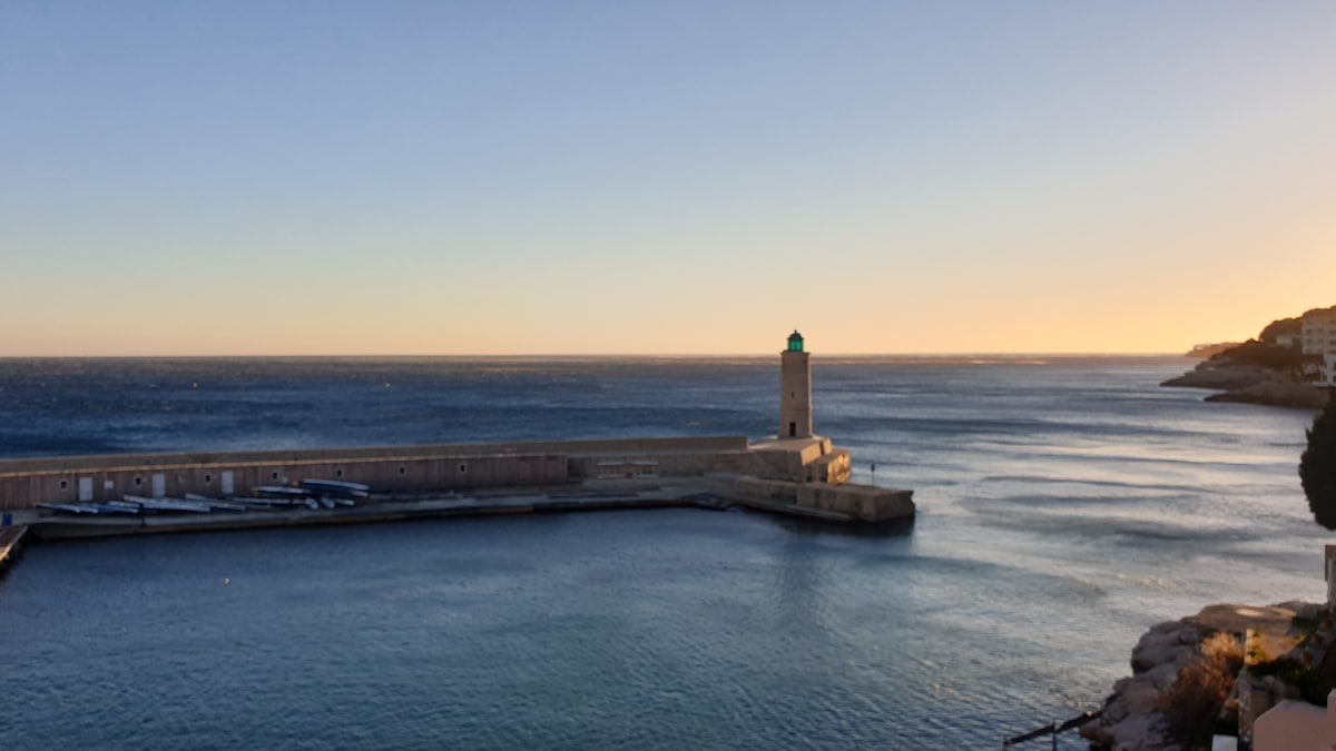 A coastal view features a lighthouse standing on the edge of a harbor, with calm waters reflecting the soft glow of the setting sun. The shoreline curves gently, and fishing boats are moored near the waterfront, framed by the serene backdrop of the horizon.