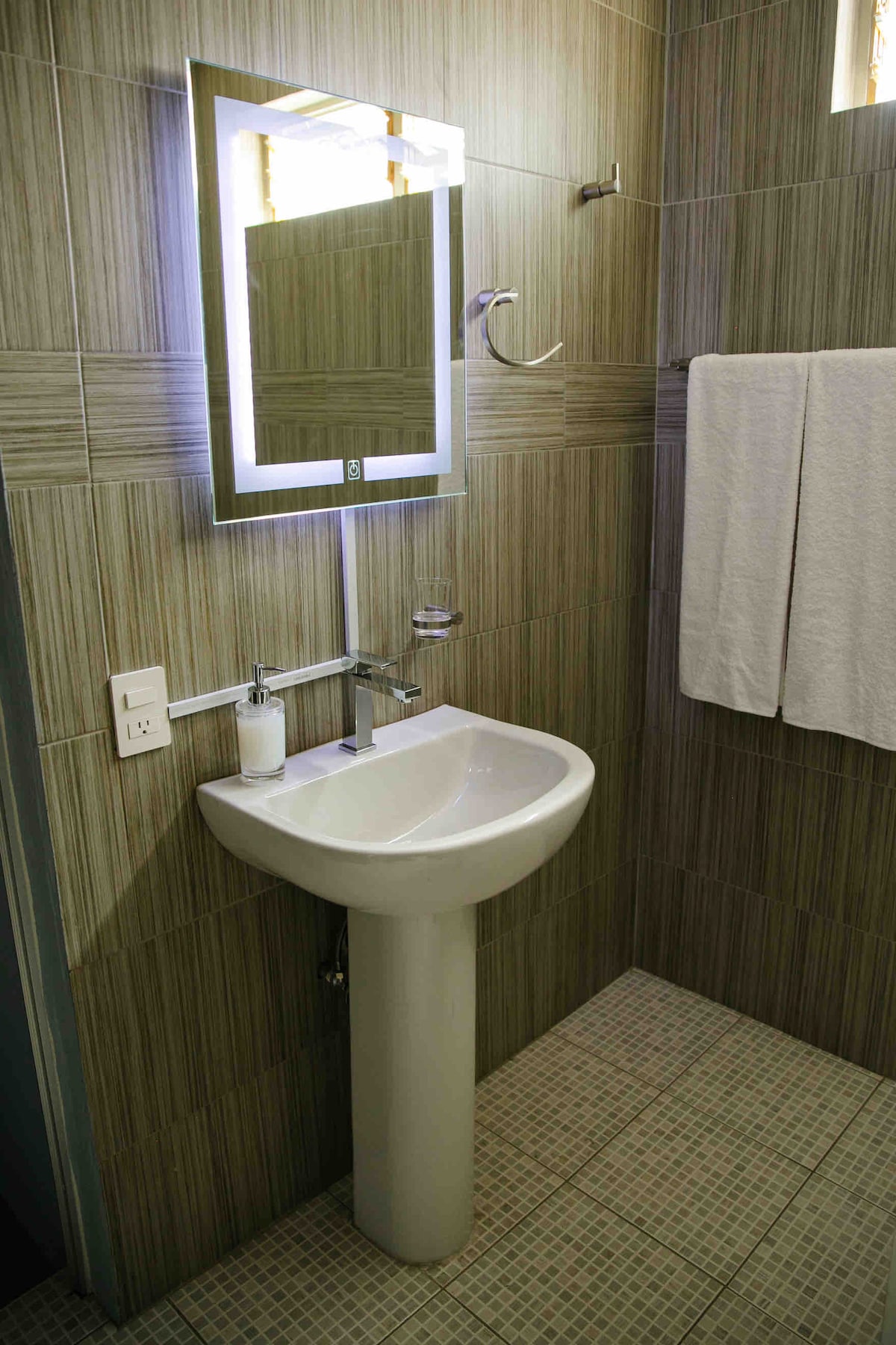 A modern bathroom space features a wall-mounted mirror with integrated lighting above a white sink on a sleek pedestal. Neutral-colored tiles line the walls and floor, while fresh white towels hang neatly on a towel rack, contributing to a clean and functional design.