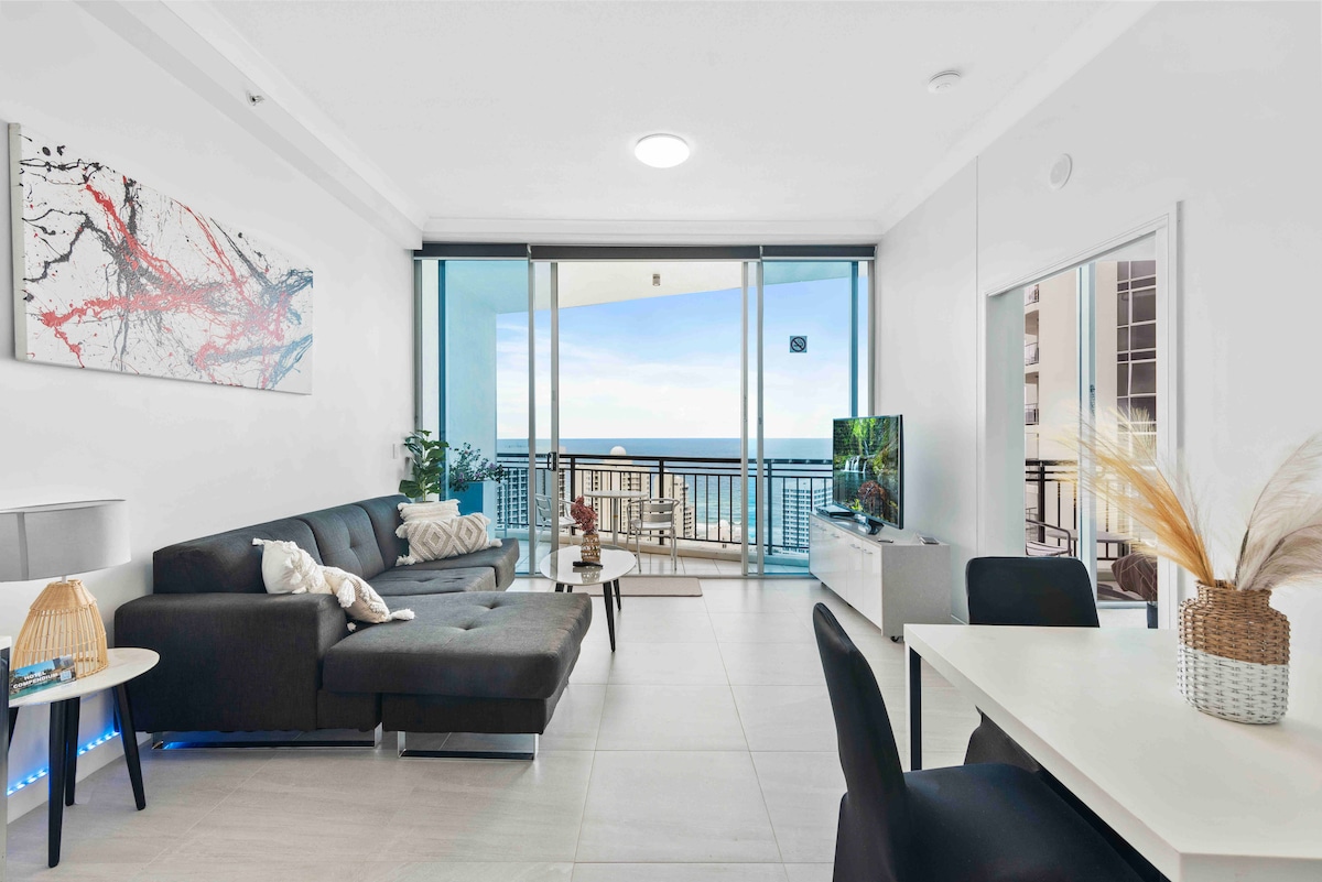 The living area features a modern black sectional sofa and a glass coffee table. A TV is positioned on a white console. Large sliding doors provide access to a balcony with ocean views, allowing natural light to fill the space. A dining table is visible in the background.