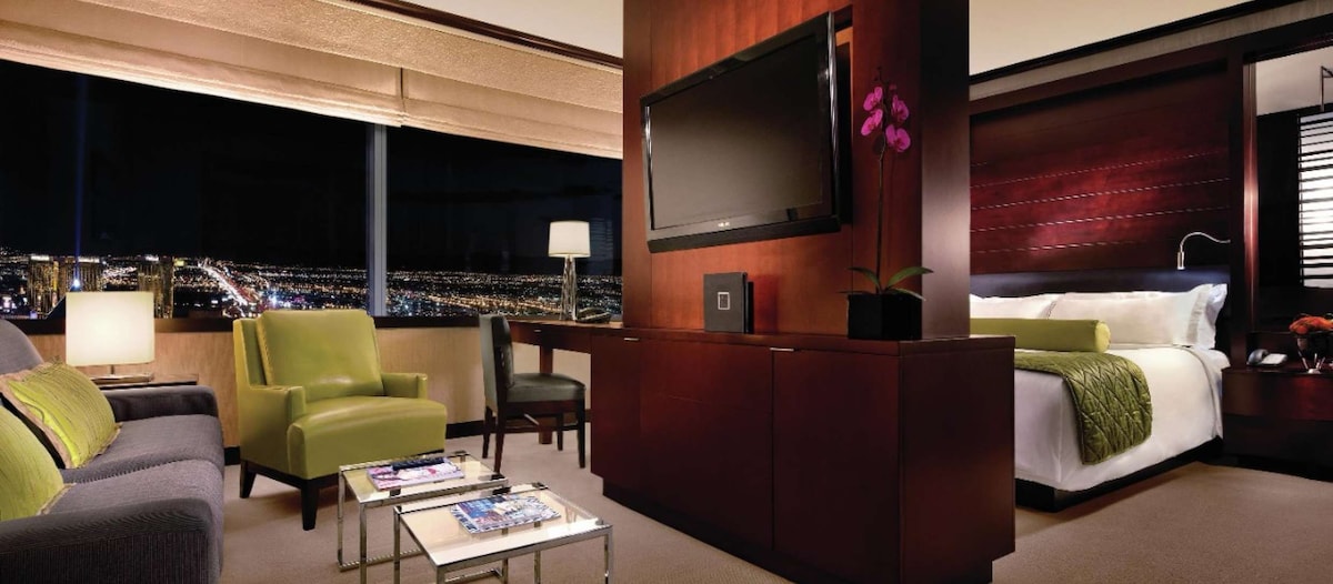 A contemporary living space is highlighted by a large flat-screen TV mounted on dark wood cabinetry. A sleek coffee table sits in front of a plush sofa and accent chair, while soft lighting creates a cozy ambiance. A king bed is visible beyond, with city lights twinkling through the window.