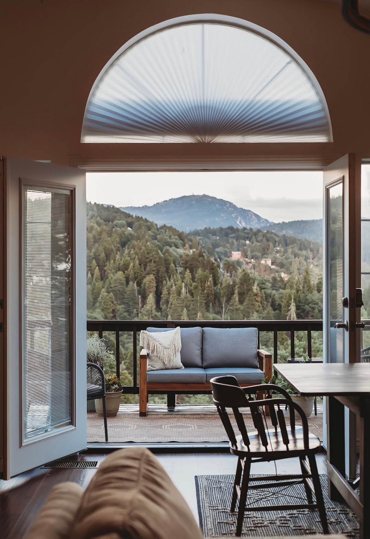 French doors open to a furnished deck, revealing a stunning mountain view. A comfortable seating area features a cozy two-seat couch, while lush greenery complements the serene landscape, creating a calming focal point just outside.
