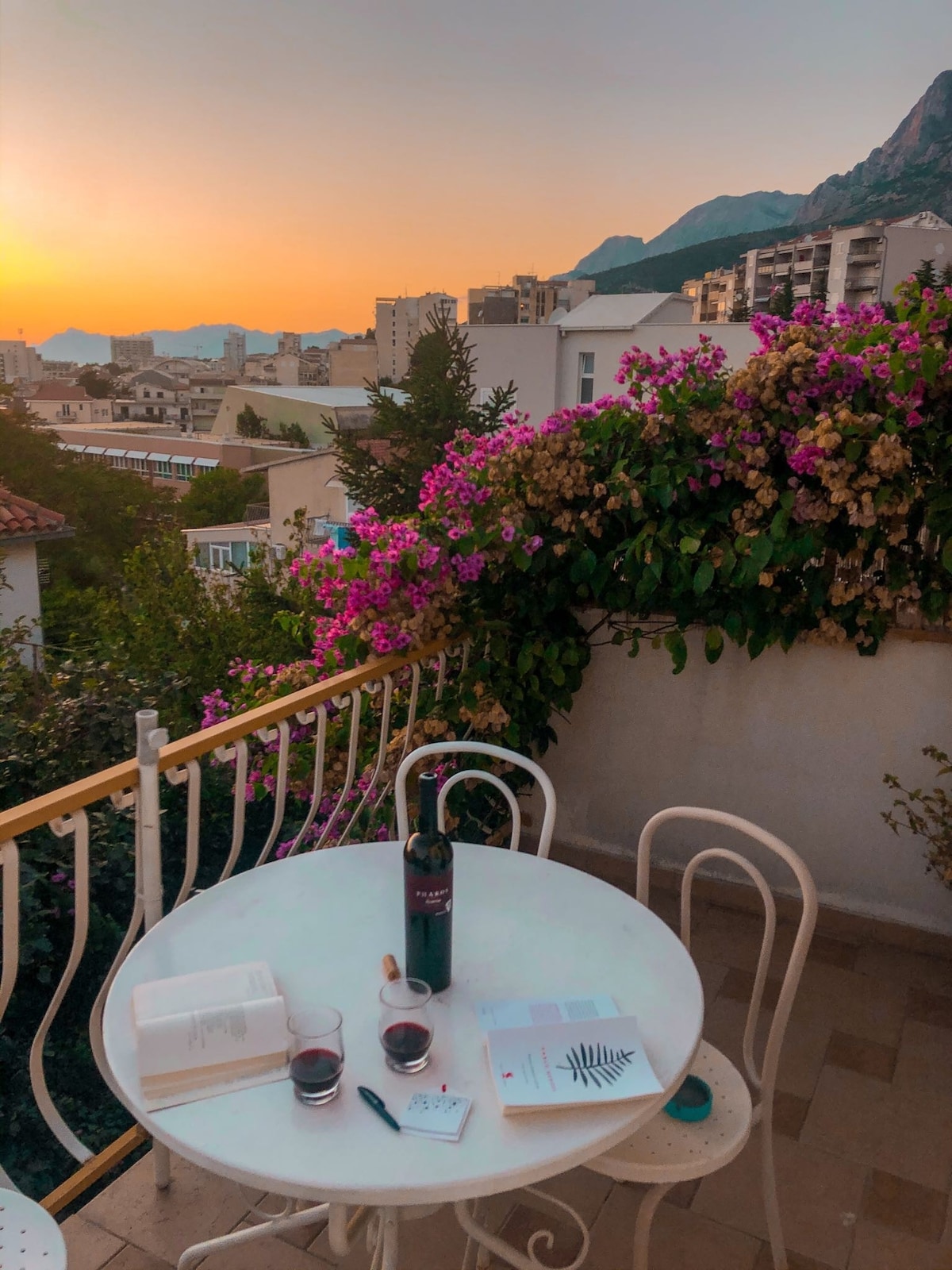 A small balcony is framed by vibrant bougainvillea, offering a view of the sunset over the city. A round table is set with a bottle of wine, two glasses, and an open book, inviting moments of relaxation amidst a serene atmosphere.