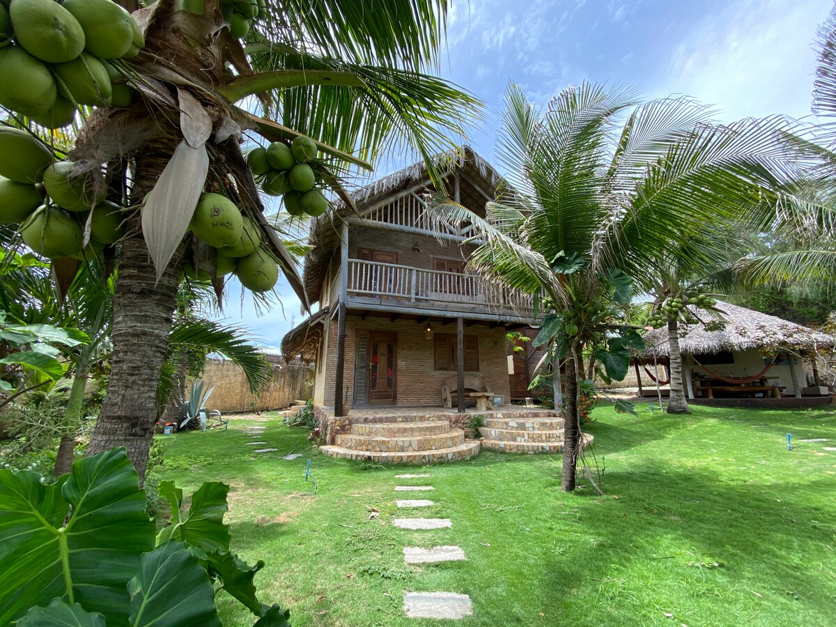 A rustic two-story house is framed by palm trees and lush greenery. A path of stepping stones leads to a welcoming entrance, lined with a spacious porch that overlooks the garden. The property features well-maintained grass and tropical plants, enhancing the outdoor ambiance.