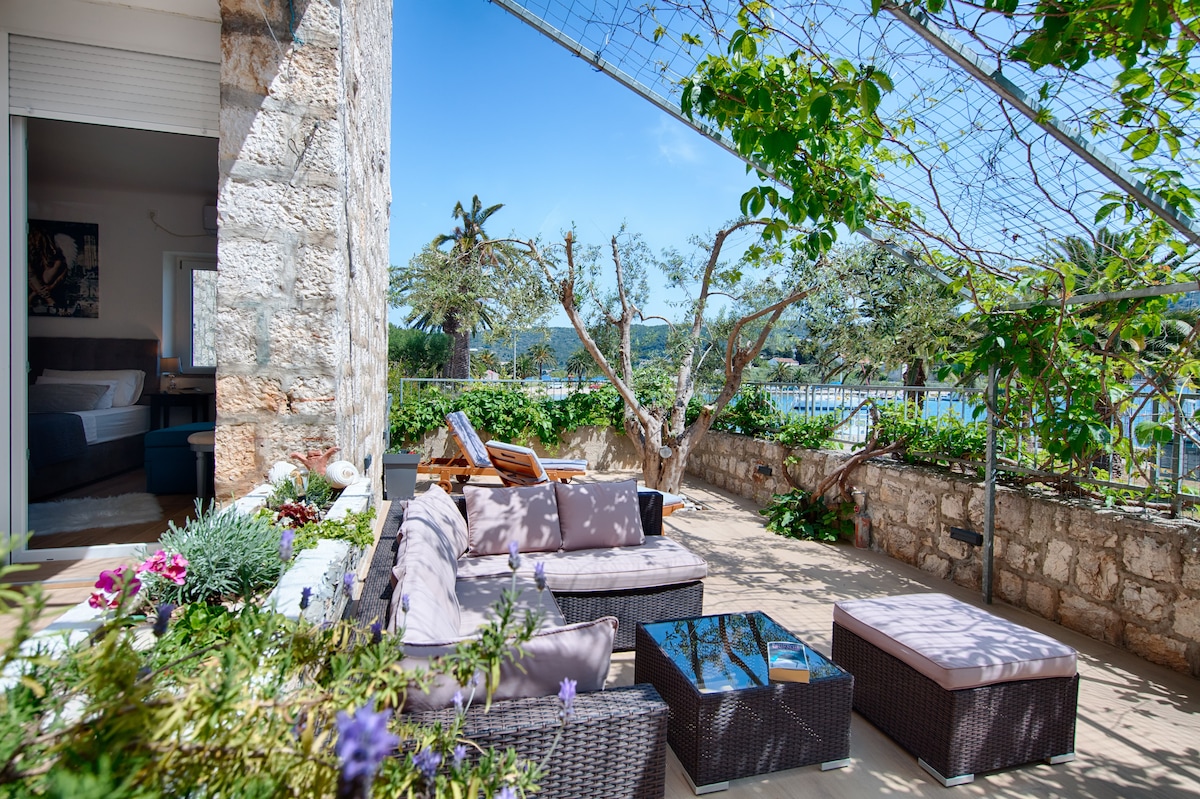 A spacious terrace is framed by lush greenery, featuring comfortable seating with plush cushions. A coffee table is positioned in the center, surrounded by vibrant plants. The area is bathed in natural light, creating a serene outdoor retreat.