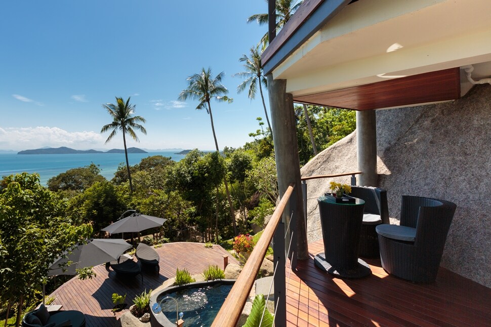 An outdoor balcony offers serene views of the coastline, surrounded by lush tropical greenery and palm trees. Two stylish chairs are positioned near a small table, with a glimpse of the plunge pool and sun loungers visible below, inviting relaxation in a peaceful setting.