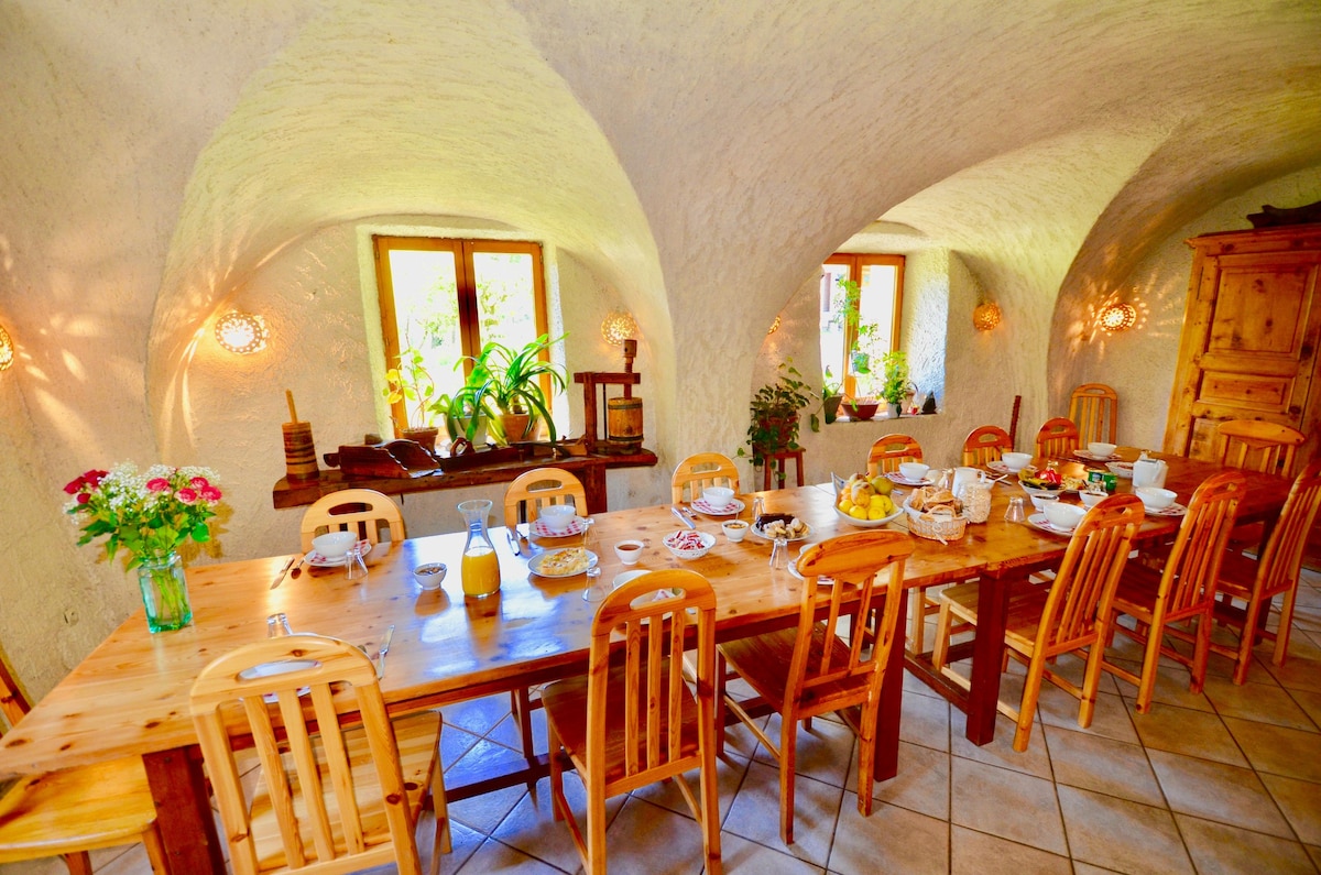 A spacious dining area is featured with a long wooden table surrounded by wooden chairs. Natural light filters through the windows, illuminating the room's arched ceilings and soft wall textures. Decorative plants and a wooden storage unit add warmth to the inviting space.